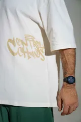 Remera oversize de algodón color off-white, con mangas cortas amplias y cuello redondo. Presenta un pequeño estampado en el centro del pecho y un diseño gráfico más grande en el lateral inferior.