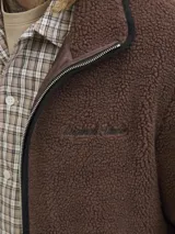 Campera de tejido tipo teddy color marrón, con cuello alto, cierre frontal completo y bolsillos laterales. Presenta un diseño de textura suave y abrigada con detalles de ribetes en color negro.