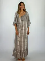 Vestido largo de corte holgado con estampado tie-dye en tonos grisáceos. Presenta escote redondo con cordones ajustables y borlas, mangas tres cuartos con volados y ruedo con volado amplio.