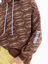 Buzo marrón con capucha y cordones en contraste, de corte amplio y estampado integral de labios. Cuenta con mangas largas y detalle de logo estampado en una de las mangas.