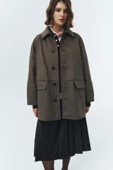 Chaqueta de gamuza sintética color verde militar, con cuello camisero y cierre frontal de botones. Presenta un diseño de corte amplio con bolsillos delanteros de solapa y mangas largas. El material tipo suede ofrece una textura suave y un estilo clásico de inspiración masculina.