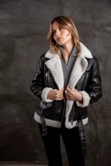 Campera tipo gamulán de eco cuero negro con interior y solapas de corderito sintético en color crudo. Presenta cierre frontal, bolsillos laterales con cierre y cinturón ajustable en la cintura.