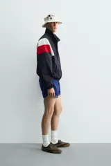 Chaqueta técnica de corte relajado con cuello subido y cierre frontal de cremallera bidireccional. Presenta un diseño color block en tonos azul marino, blanco, rojo y azul eléctrico. Incluye bolsillos frontales de vivo, puños y ruedo elásticos, y forro interior de red.