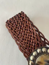 Cinto ancho de cuero trenzado en color marrón, con hebilla circular decorativa que presenta un centro de piedra natural y borde con apliques metálicos esféricos.