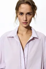 Camisa de algodón color lila, con cuello camisero ribeteado en encaje, cierre frontal de botones con detalles de encaje a los lados y mangas globo largas con puños elásticos.