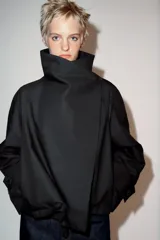Cazadora corta de color negro con cuello subido cruzado, manga larga con puños anchos ajustados con trabilla y botón, y cierre frontal cruzado con botones ocultos.
