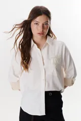 Camisa blanca de manga larga con cuello clásico, cierre frontal con botones y bolsillo en el pecho con bordado de una planta.