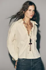 Collar largo con cadena de eslabones dorados y cuentas redondas en tonos rojo oscuro y verde azulado. El dije principal es una pieza metálica dorada con relieve de un pájaro, adornado con pequeñas piedras de colores, y un colgante de borla de cadena metálica oscura, unido a una pieza rectangular de resina color borgoña.