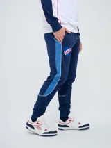 Pantalón de jogging azul marino Umbro del Club Nacional de Football, con cordón ajustable en la cintura, puños elastizados en los tobillos y bandas laterales en tonos de azul. Cuenta con el escudo del club en una pierna y el logo de la marca en la otra.