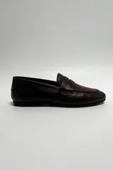 Mocasín de cuero color café, diseño clásico de estilo penny loafer con suela plana y costuras visibles.