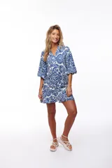 Vestido corto blanco con estampado floral azul, escote en V, mangas 3/4 con volados y falda con volados. Cierre con botones en el frente.