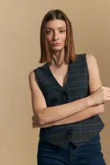 Chaleco sastrero de corte entallado con escote en V y cierre frontal de botones. Presenta un diseño de estampado a cuadros en tonos oscuros con líneas finas en azul y naranja. Incluye terminación en punta en el ruedo y ajuste con hebilla en la espalda.