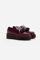 Zapato tipo mocasín de plataforma color bordó, confeccionado en una combinación de cuero liso y gamuza. Presenta cordones bicolores con detalle de borlas decorativas en el empeine.