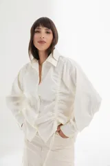 Camisa blanca de mangas largas con cuello clásico y cierre con botones. Presenta detalles de fruncido vertical en la parte delantera y cordones ajustables en los laterales. Corte holgado.