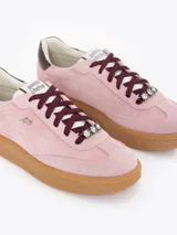 Zapatilla de cuero descarne color rosa, con suela de caucho color caramelo. Presenta cordones oscuros con un aplique de cristal decorativo y detalle de cuero liso en el talón.