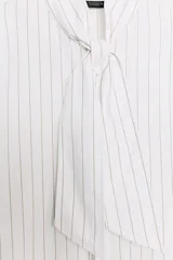 Camisa de popelín de algodón a rayas verticales, con cuello subido y aplicación de tejido tipo pañuelo anudado al frente. Presenta manga larga con puños anchos y cierre frontal con botones ocultos.