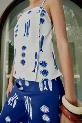 Top de escote recto con tirantes finos ajustables, estampado con motivos marinos de peces y conchas en color azul sobre fondo blanco. Presenta detalle de botones decorativos en el frente y aberturas en el bajo.
