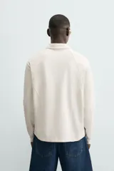 Polo de manga larga color crema, confeccionado en tejido de algodón, con cuello solapa y cierre frontal de botonadura. Presenta un calce relajado y acabados en rib en puños y ruedo.