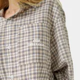 Camisa de manga larga con corte boxy fit, confeccionada en tela a cuadros en tonos beige y marrón. Presenta cuello clásico, cierre frontal con botones y un bolsillo en el pecho con el logo de la marca bordado.