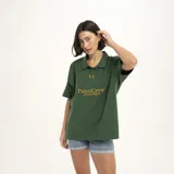 Remera tipo polo de manga corta, color verde oscuro, con cuello y abertura frontal. Presenta un estampado dorado en el pecho con el texto "PulauCrew" y un pequeño logo de corona de laurel bordado.