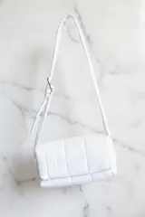 Cartera de hombro acolchada color blanco, con diseño de cuadrícula y correa ajustable.