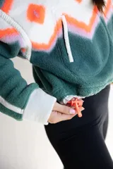 Buzo de polar con diseño estampado de estilo étnico en tonos verde, blanco y naranja. Presenta cuello alto con cierre frontal parcial, mangas largas con puños en contraste y cordón ajustable en el ruedo.