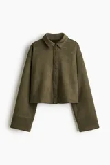 Camisa corta color verde militar, de corte holgado, con cuello clásico, botones frontales y canesú en la espalda. Presenta hombros caídos y mangas largas con puños anchos.