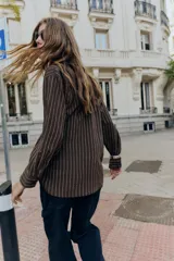 Camisa de cuello solapa y manga larga, color marrón con rayas verticales blancas. Presenta cierre frontal con botones ocultos y dos hebillas decorativas en color beige en el lateral.