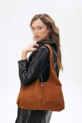 Cartera tipo hobo de cuero ovino color suela, con diseño amplio y flexible. Presenta un aplique metálico pequeño en el frente y costura central vertical.