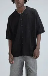 Camisa de manga corta tejida en punto abierto tipo crochet, color negro, con cuello polo y cierre frontal de botones. Presenta un diseño de calce oversize.