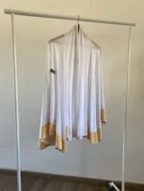Ruana de tela fina con diseño en color blanco y detalles en amarillo en el ruedo. Presenta una caída suave y un parche bordado rectangular en la parte superior.