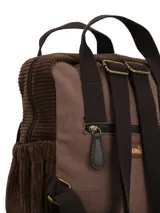 Mochila de diseño patchwork en tonos marrones, confeccionada en textil con detalles de pana y gamuza. Cuenta con manijas superiores fijas, bolsillo frontal con solapa, compartimento principal con cierre y un llavero decorativo con borlas y cuentas colgando de un lateral.