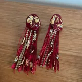Caravanas colgantes con base de resina color bordó con incrustaciones de láminas doradas y cristales, acompañadas de flecos de hilo rojo y cadenas de strass.