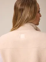 Chaleco de sherpa color beige con cierre frontal completo, cuello alto y bolsillo con cierre en el pecho que incluye un detalle circular con logo.