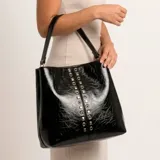 Cartera al hombro Korium modelo Anthea, color negro con efecto charol arrugado y detalles de tachas y argollas metálicas plateadas en la parte frontal.