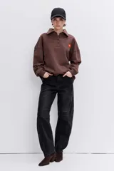 Sudadera de algodón color marrón, con cuello polo y cierre frontal de botones. Presenta un diseño holgado, mangas largas con puños elásticos y un pequeño parche bordado en forma de corazón en el pecho.