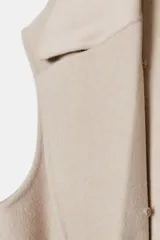 Chaleco largo de mezcla de lana color beige, de corte recto con manga sisa. Cuenta con cuello solapa con aplicación de pelo sintético extraíble, bolsillos laterales ocultos y un cinturón ajustable del mismo tejido para anudar en la cintura.