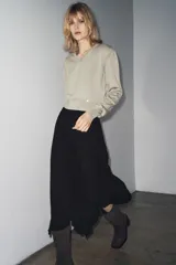 Jersey de punto color beige claro, con cuello en V y manga larga. Presenta detalles de botones decorativos en los hombros y en el ruedo inferior, sugiriendo un diseño desmontable o con aberturas ajustables.