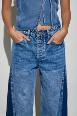 Jean de denim en tonos azul claro y azul oscuro, de tiro alto y corte wide leg, con inserciones laterales en contraste, detalles de tachas metálicas y acabado desmechado en el ruedo.
