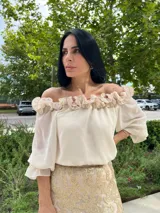 Blusa color beige claro, estilo campesino, con escote Bardot (hombros descubiertos) adornado con volados fruncidos. Mangas largas abullonadas con puños elásticos.