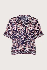 Camisa de manga corta con cuello solapa y cierre frontal de botones. Presenta un estampado geométrico tipo ikat en tonos azul marino, blanco y detalles en rojo, con un borde decorativo en el ruedo y las mangas.