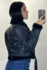 Campera tipo aviador de cuero sintético negro con interior y detalles de piel sintética al tono en cuello, puños y ruedo. Presenta cierre frontal cruzado y cinturón ajustable con hebilla metálica en la cintura.