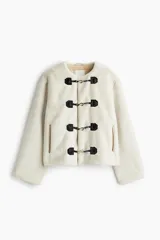 Campera de tejido suave efecto peluche en color crudo, con cuello redondo, hombros caídos y cierre frontal mediante alamares con detalles en contraste negro. Cuenta con bolsillos laterales ribeteados y forro interior.