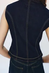 Chaleco de denim azul oscuro con cuello redondo y cierre frontal de botones. Presenta un diseño de pespuntes decorativos en contraste con forma de alamares en la parte delantera.