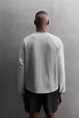 Camiseta de algodón gris de corte relajado con mangas largas ranglán de color gris más claro y puños elásticos. Presenta un pequeño bordado en el pecho y un parche con logo en la parte inferior delantera.