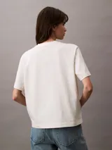 Remera de algodón color blanco hueso, corte boxy, con mangas cortas con dobladillo y logo de Calvin Klein estampado en verde. Diseño de estilo universitario.
