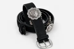 Cinto negro de cuero sintético con apliques metálicos circulares grabados con motivos florales y hebilla metálica de estilo rústico.