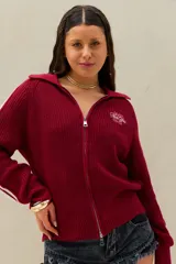 Sweater de punto color bordó con cierre frontal completo de doble sentido, cuello tipo polo y manga larga con detalles de líneas blancas en el lateral. Presenta un bordado en el pecho con texto en tipografía cursiva.