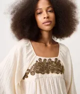 Blusa tipo bata color off-white de viscosa y poliamida, con cuello redondo, mangas cortas amplias, fruncido bajo el busto y bordado delantero contrastante.