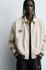 Cazadora estilo varsity de tejido efecto ante en color beige. Presenta cuello solapa, cierre frontal con botones a presión y puños y ruedo con acabado en rib a rayas negras. Incluye parches bordados decorativos en el pecho y la manga, además de bolsillos laterales de doble vivo.